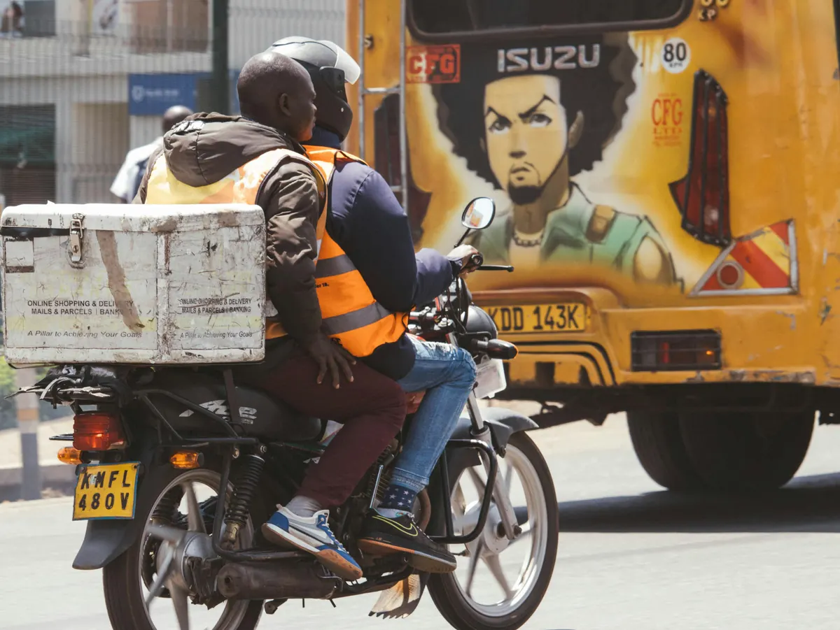 Two people riding a motorcycle past a brightly painted bus in an African city