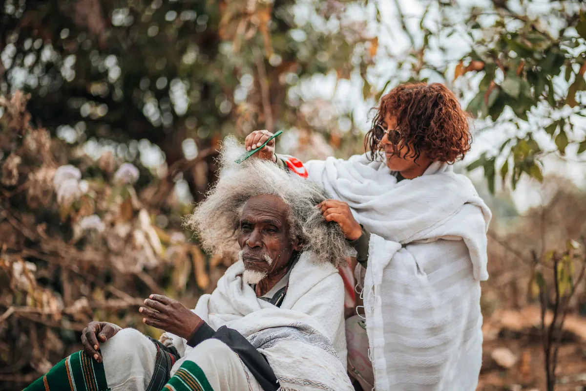 A woman braiding an older man's hair outdoors
