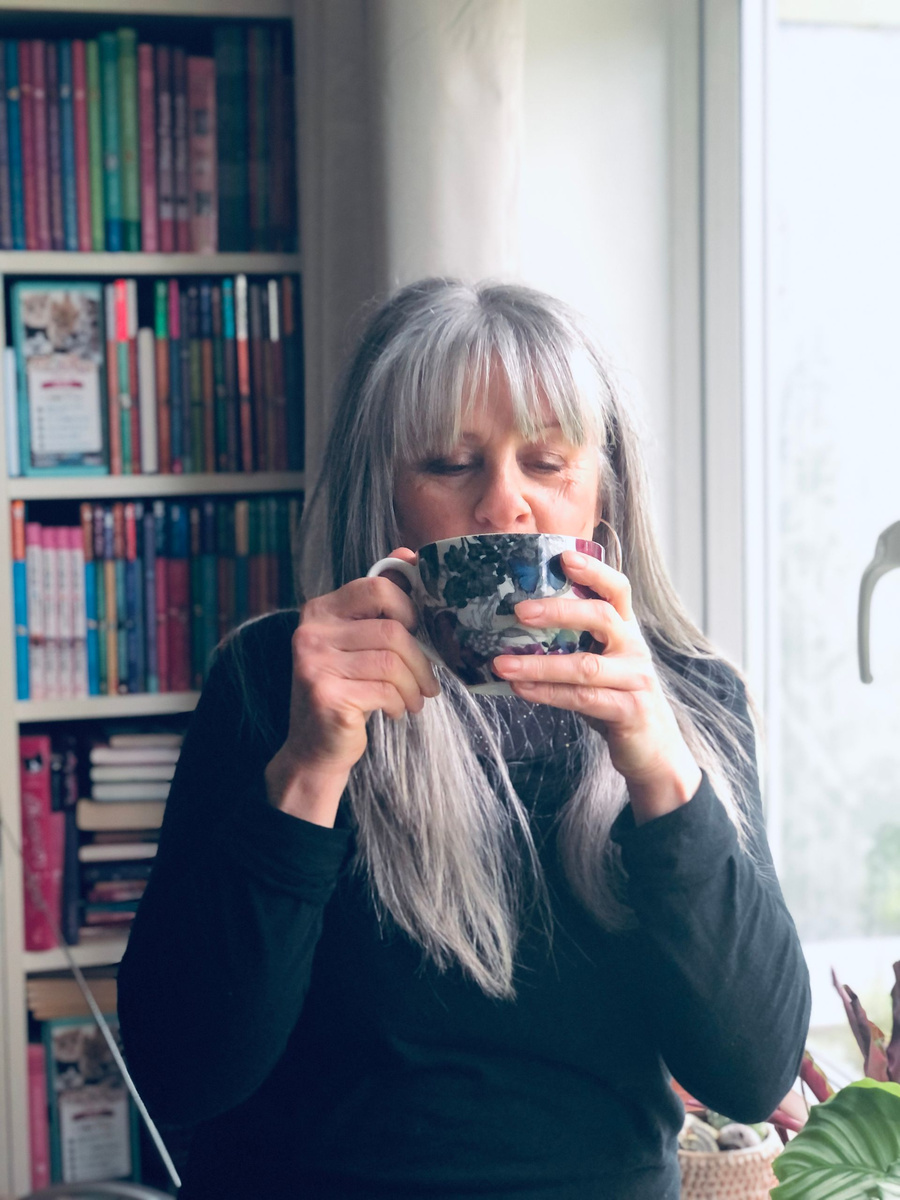 Sue Bentley drinking from a mug beside her bookshelves