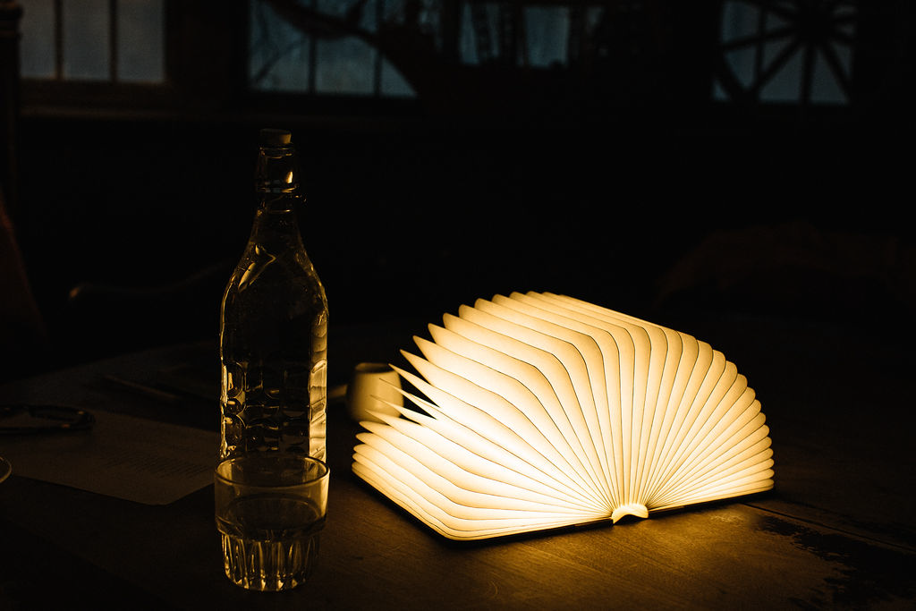 A glowing open-book lamp sits on a dark table beside a glass bottle and tumbler, creating a moody, atmospheric reading scene.