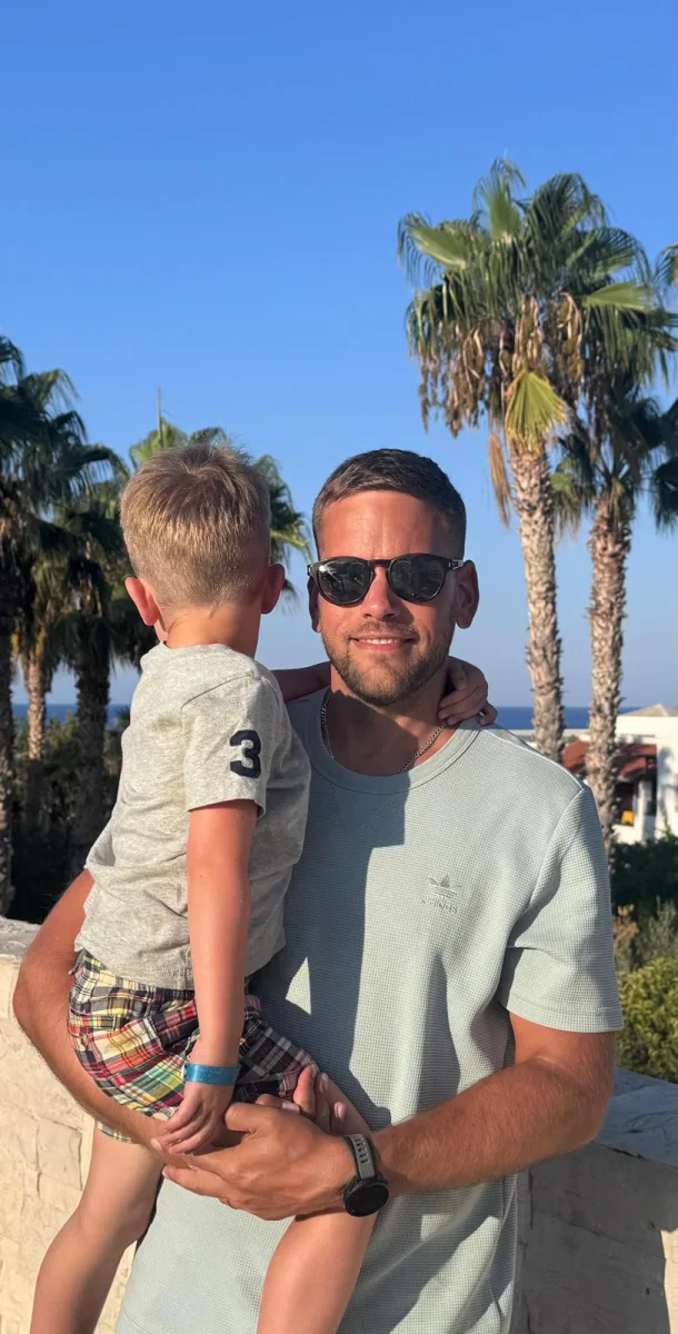 Sam Macadam outdoors, holding a young child beneath palm trees.