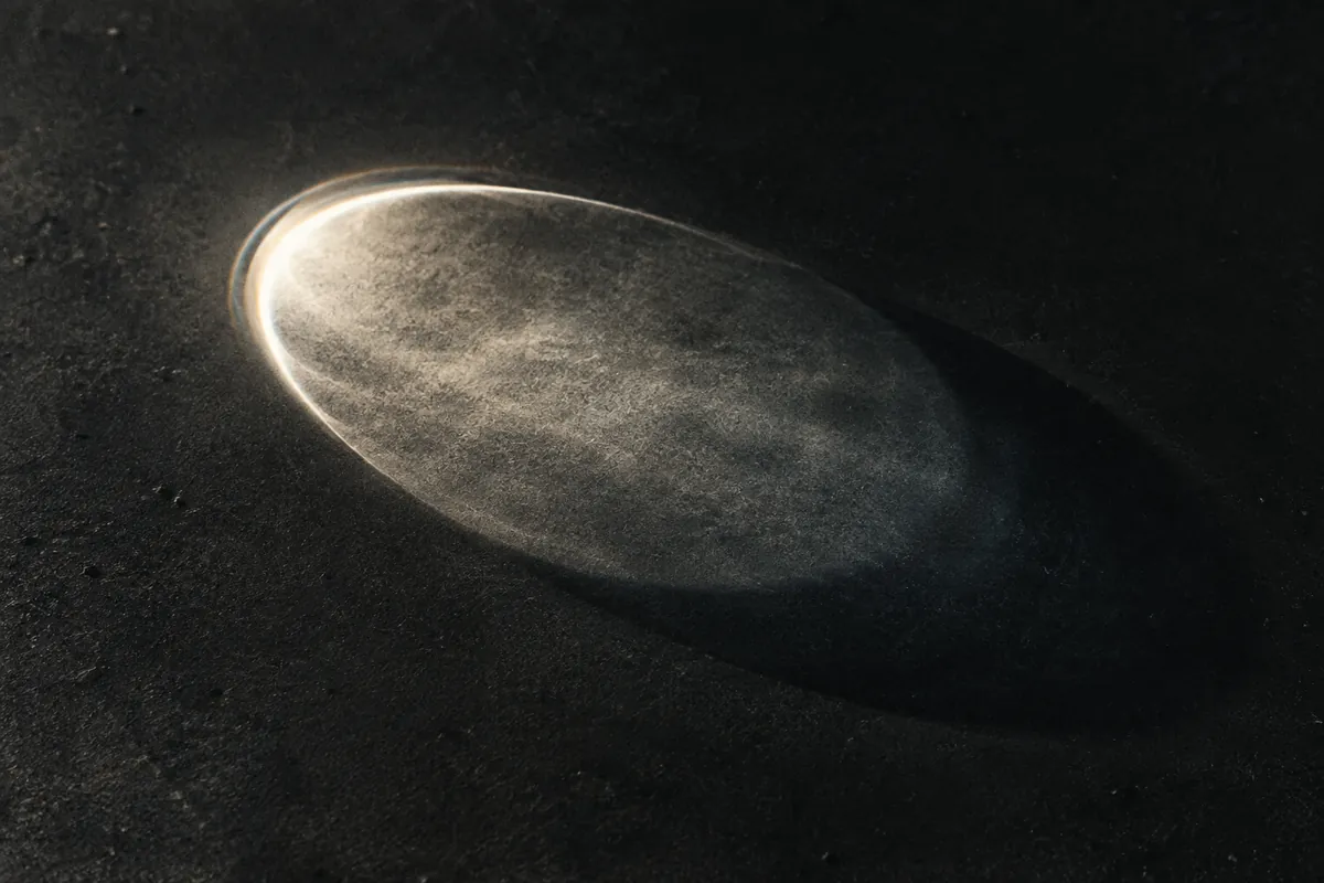 An oval of refracted ivory light beside a soft shadow on a charcoal surface.